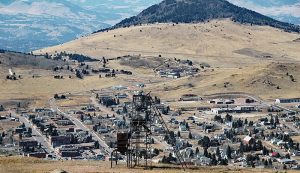 Photo shows an aerial view of Cripple Creek Colorado. Yellow grasses surround the town on hills, and a large mountain stands over the small town.